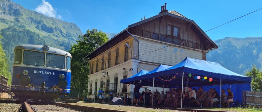 Ein blauer Zug steht auf der Eisenbahnschiene. Ein großes Gebäude und ein Zelt befinden sich neben dem Zug. Viele Menschen sitzen unter dem Zelt.
