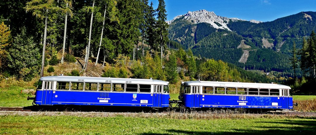 Ein blauer und weißer Zug ist auf den Gleisen geparkt, mit einem Gebirgszug im Hintergrund. Der Zug ist von Bäumen und Gras umgeben.