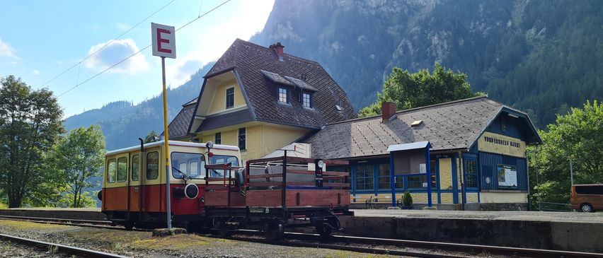 Ein kleiner Zug auf einem Gleis mit einem Bahnhofsgebäude im Hintergrund, mit Bergen in der Ferne.