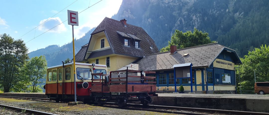 Ein kleiner Zug auf einem Gleis mit einem Bahnhofsgebäude im Hintergrund, mit Bergen in der Ferne.