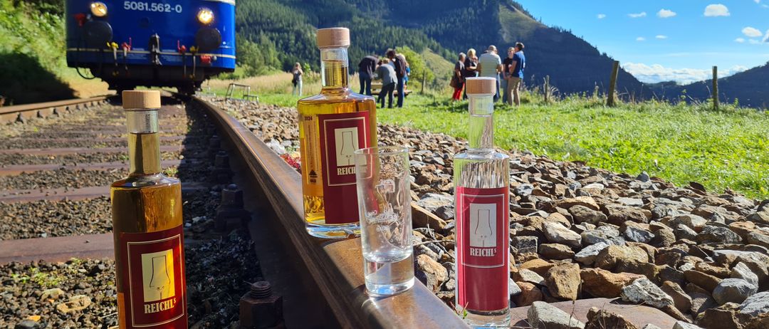 Drei Flaschen Reichl-Likör stehen auf einem Bahngleis, mit einem Zug im Hintergrund und Menschen, die auf einem Grasfeld stehen.