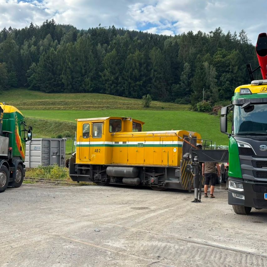A yellow and green train engine is parked on a gravel road with a green truck on the left and a red and green truck on the right.