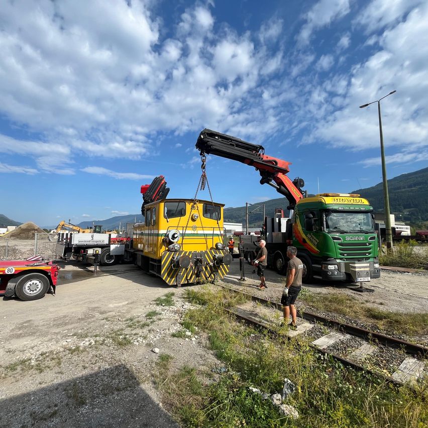 A yellow train engine is being lifted by a crane on a sunny day, with workers standing nearby.
