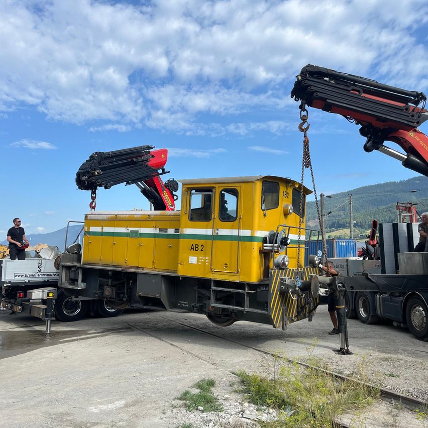 A yellow and green train car is being lifted by two cranes on a sunny day. Workers are standing nearby, and the sky is partly cloudy.