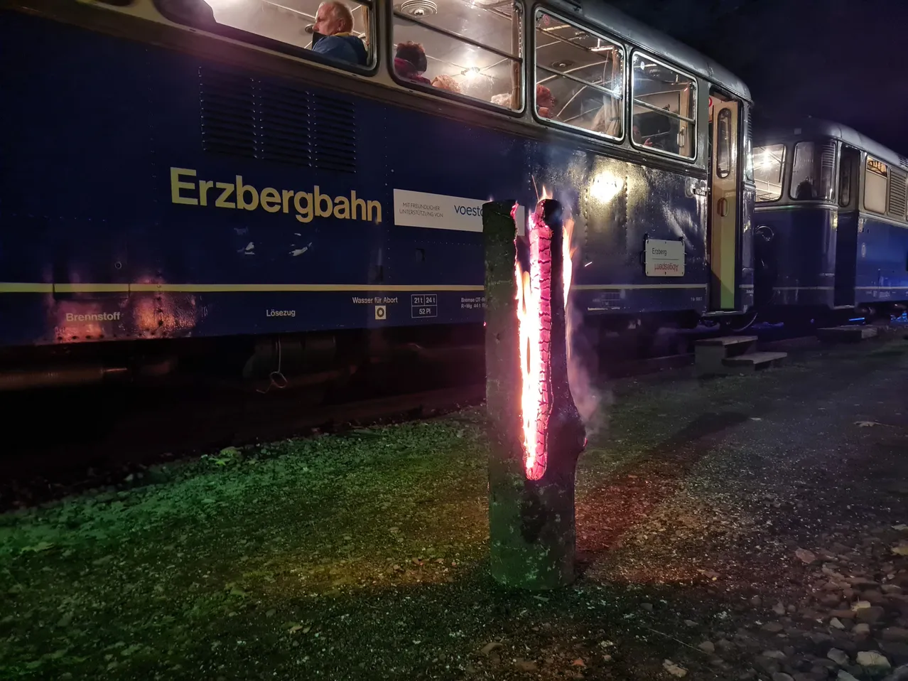 Eine Nachtszene eines blauen Zuges mit dem Wort Erzbergbahn an seiner Seite. Der Zug steht am Rande der Gleise mit einem brennenden Baumstumpf davor.