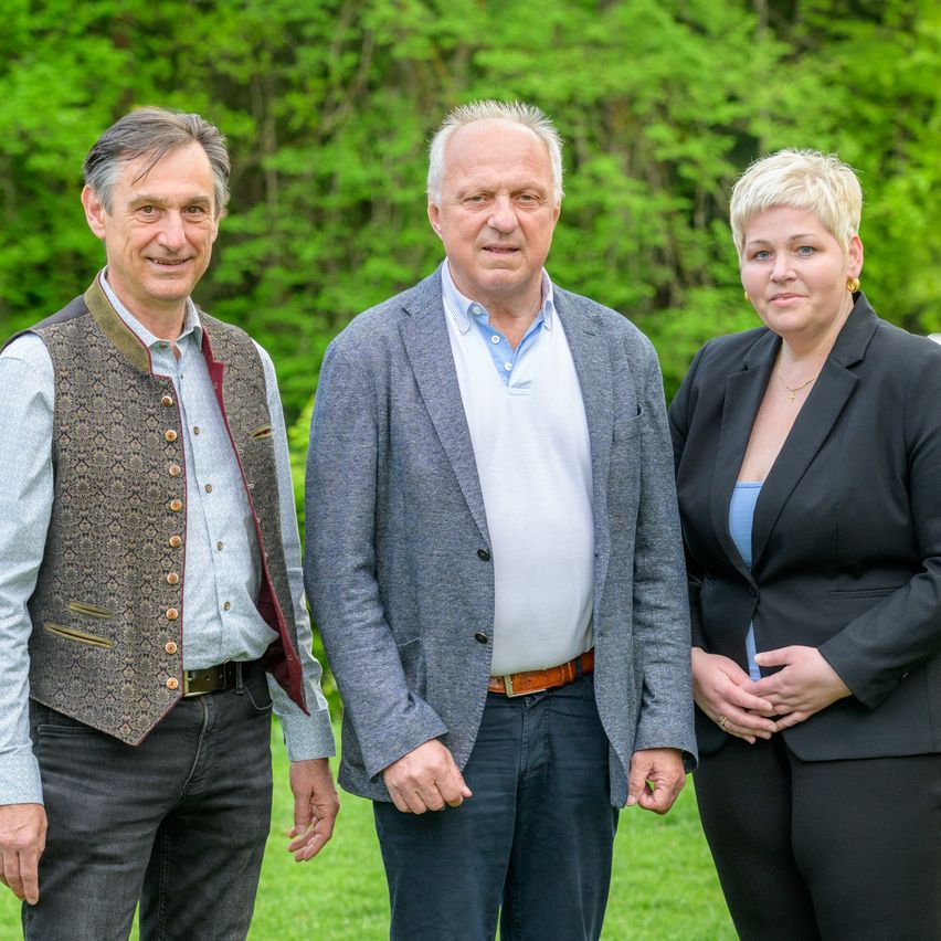 Drei Personen stehen zusammen in einem grünen Feld und posieren für ein Foto. Der Mann links trägt eine Weste, der Mann in der Mitte einen hellgrauen Anzug und die Frau rechts einen schwarzen Blazer.