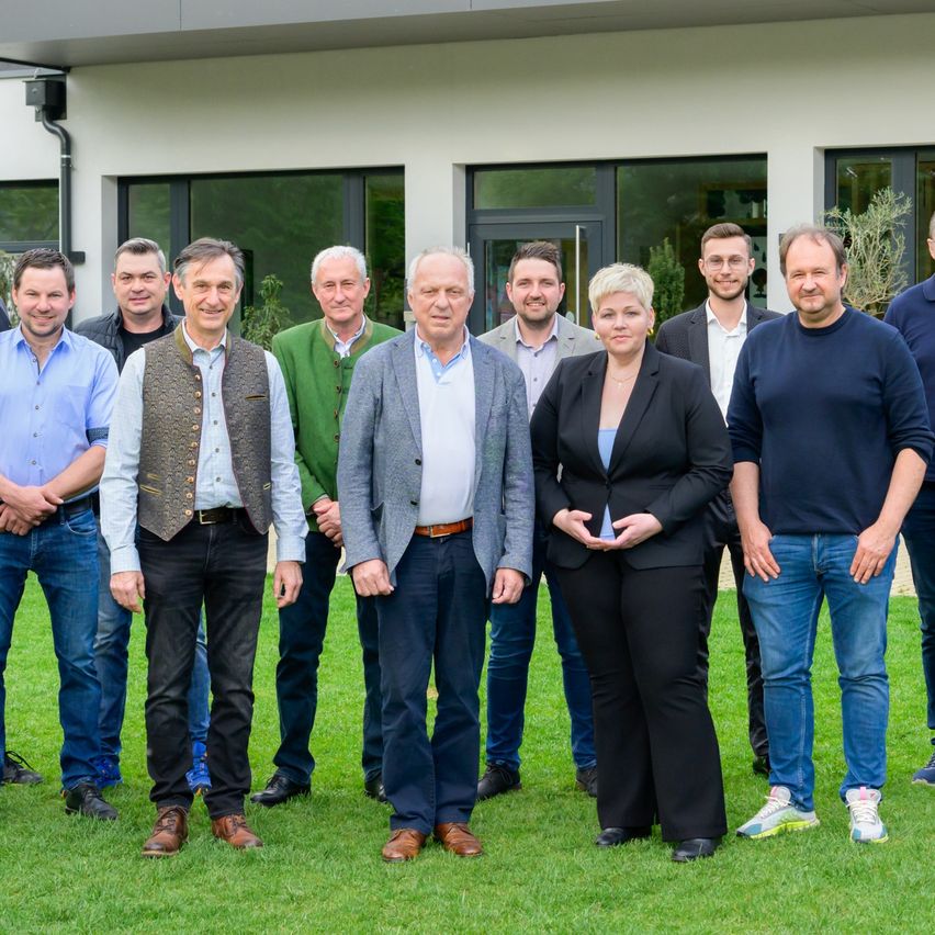Gruppenfoto eines Teams vor einem modernen Gebäude, mit Bäumen und Grünpflanzen im Hintergrund.