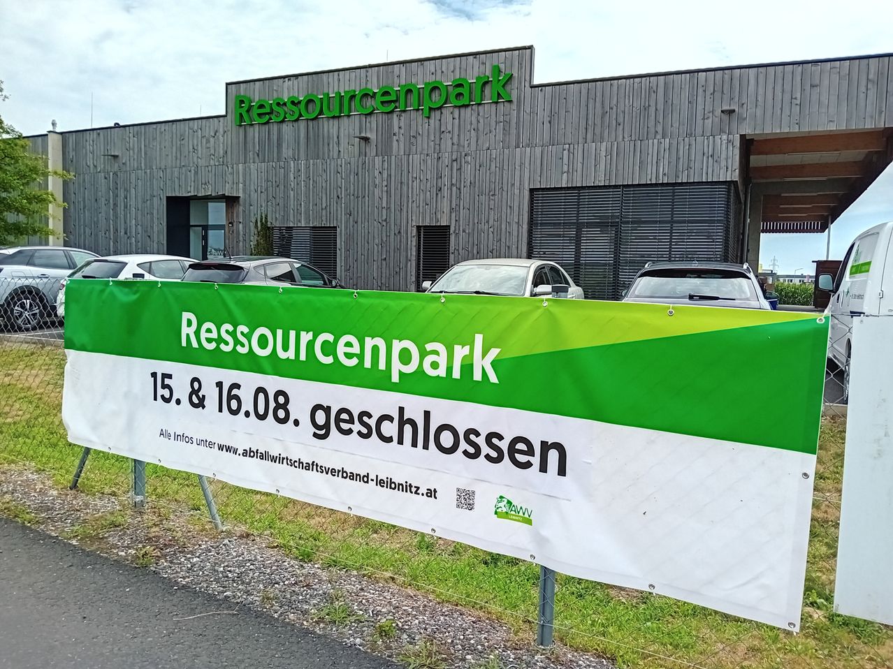 Ein Banner für Resourcenpark ist vor einem Gebäude angebracht. Es enthält das Datum eines Tag der offenen Tür am 15. und 16. August. Mehrere Autos sind vor dem Gebäude geparkt.