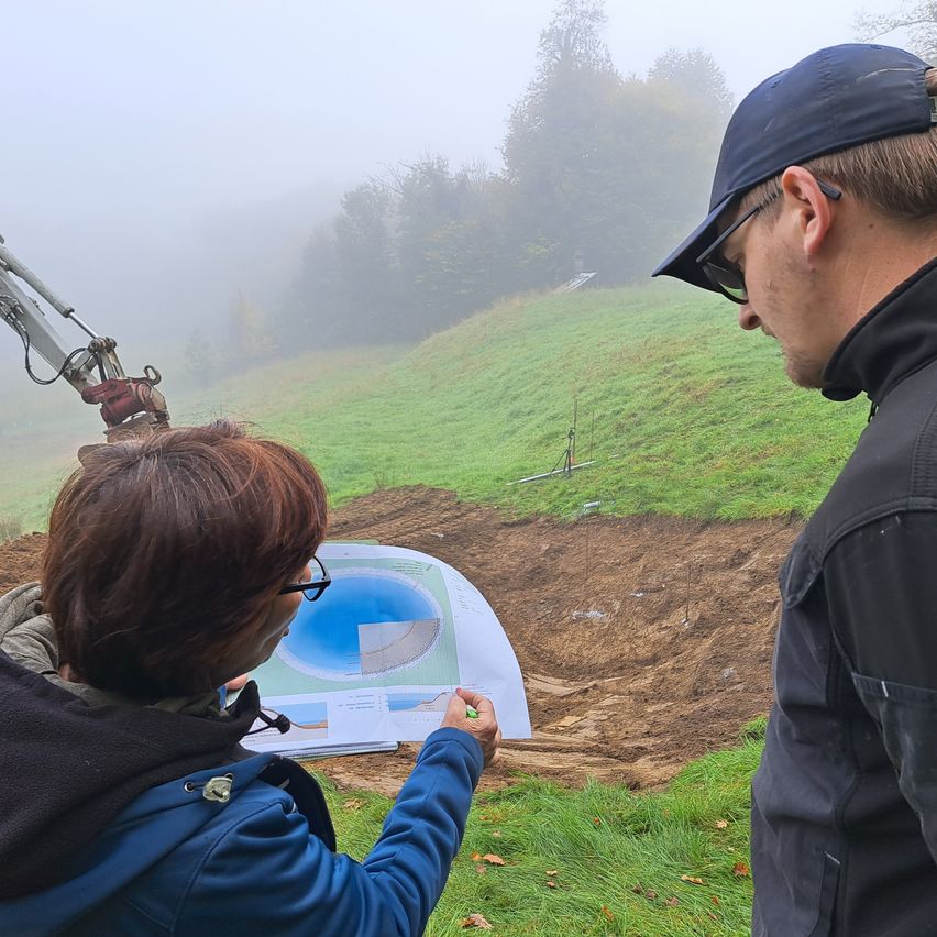 Zwei Personen auf einem nebligen Hügel, eine überprüft ein Dokument, während die andere zusieht, mit einem Baufahrzeug und einer blauen Poolzeichnung im Hintergrund.