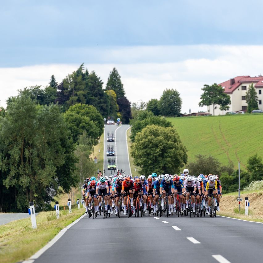 Ein Peloton von Radfahrern fährt auf einer Landstraße, mit Autos in der Ferne und Bäumen und einem Gebäude an der Seite.