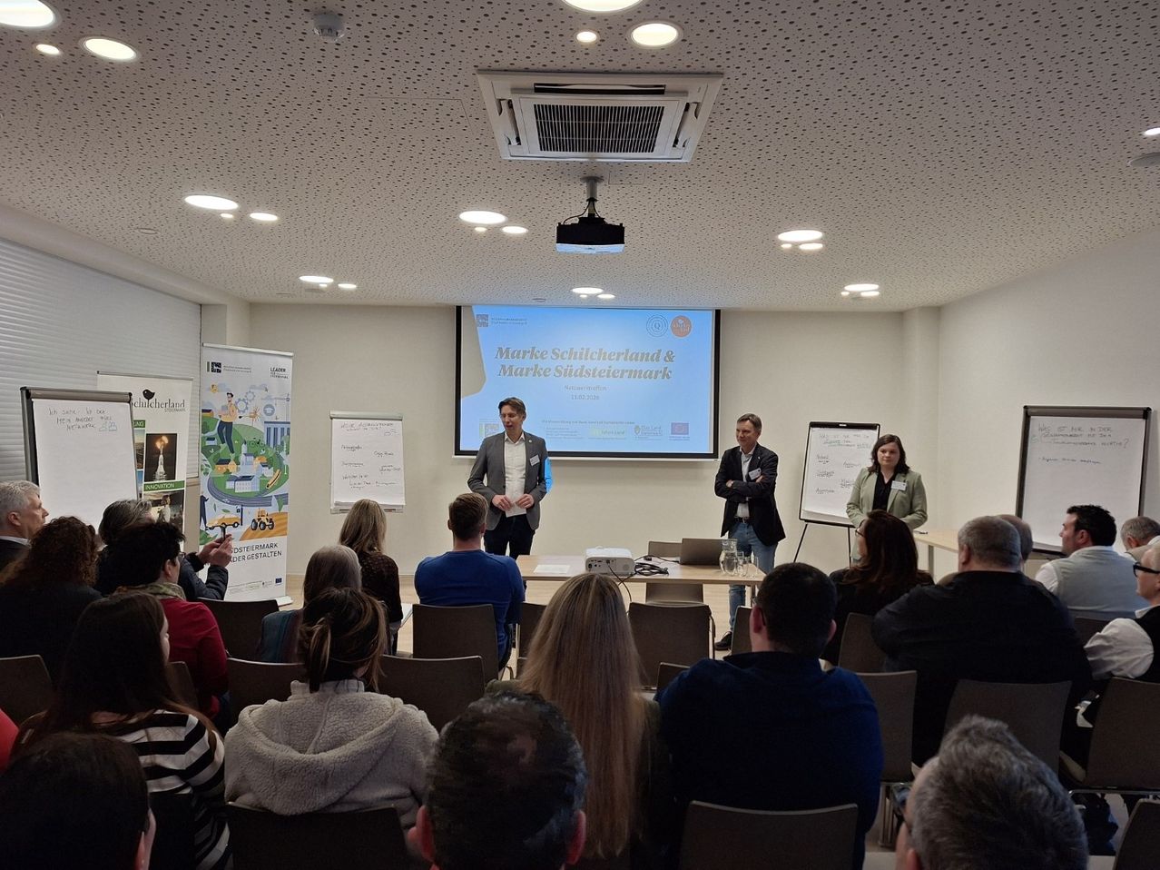 Zwei Männer und eine Frau stehen vor einer Projektionsleinwand mit einer Präsentation über Mark Schielerland und Mark Schiestermark. Das Publikum sitzt in Stühlen.