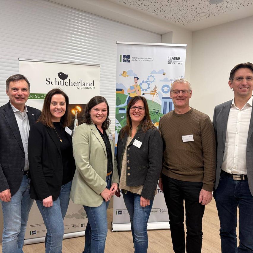 Fünf Menschen stehen nah beieinander für ein Foto und lächeln. Sie sind in einem Raum mit weißen Wänden. Hinter ihnen sind Banner mit Logos und Text.