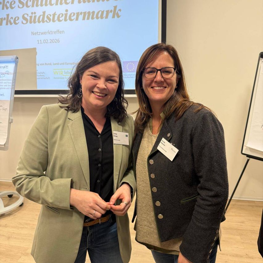 Zwei Frauen stehen in einem Raum mit Holzboden und lächeln für ein Foto. Sie tragen beide Jacken und Namensschilder. Ein Whiteboard und eine Projektionsleinwand sind im Hintergrund.
