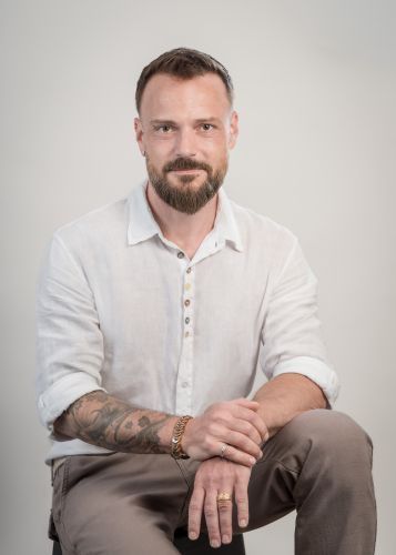 Ein Mann mit Bart, der ein weißes Hemd und braune Hosen trägt, sitzt auf einem Hocker und lächelt in die Kamera. Er hat Tattoos an den Armen und trägt eine Uhr und einen Ring.