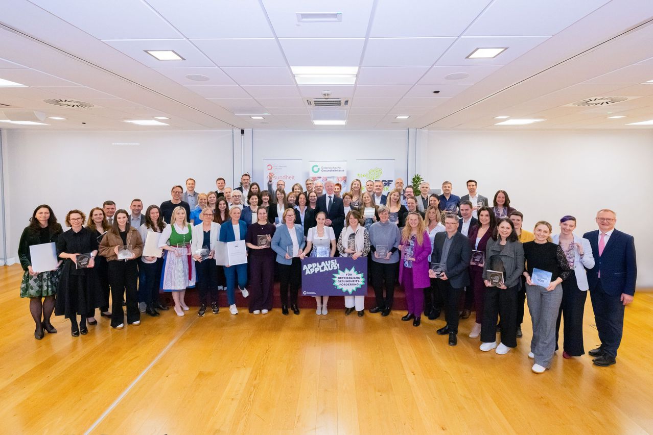 Eine Gruppe von Menschen in formeller Kleidung steht in einem Raum, einige halten Auszeichnungen. Eine lila Fahne mit weißem Text ist in der Mitte zu sehen.