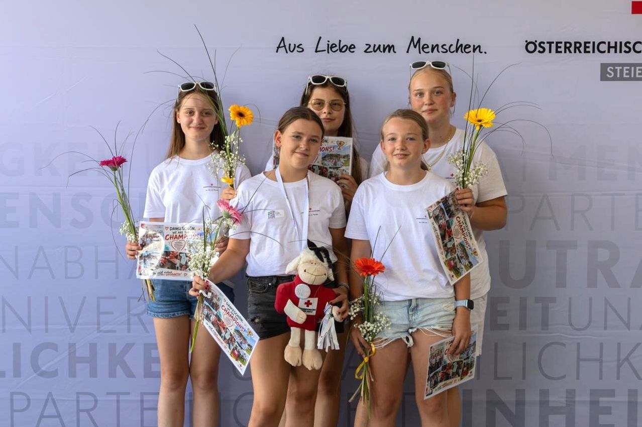 Fünf junge Mädchen stehen zusammen, halten Blumen und Urkunden und lächeln für ein Foto vor einem weißen Hintergrund mit Text.