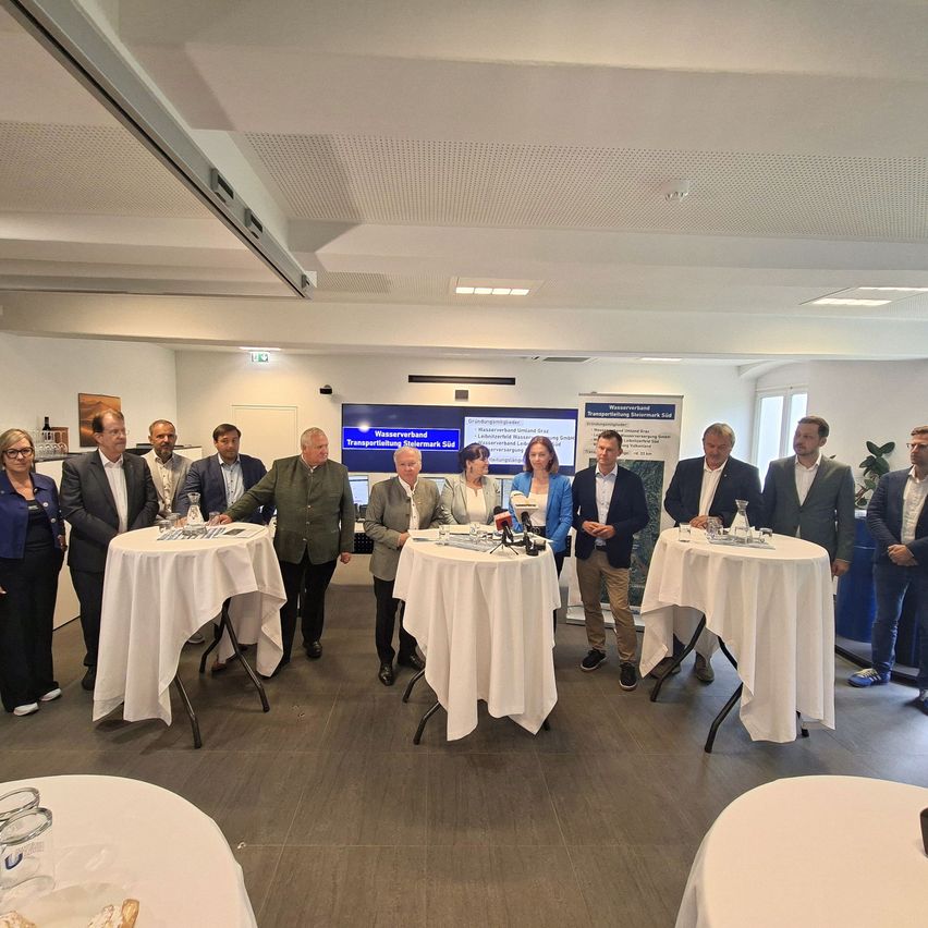 Eine Gruppe von Menschen in formeller Kleidung steht um Tische in einem Konferenzraum herum, möglicherweise um ein Projekt zu besprechen. Auf den Tischen befinden sich Mikrofone und Gläser.