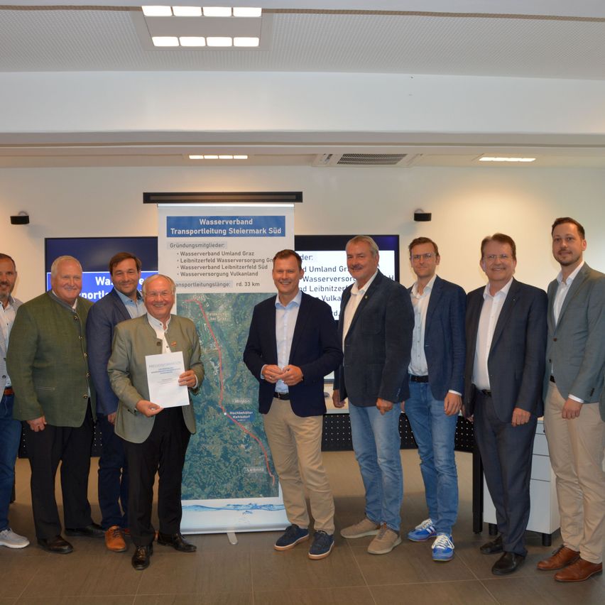 Eine Gruppe von Männern in Anzügen steht vor einer Karte und einem Banner, wahrscheinlich in einem Konferenzraum. Ein Mann hält ein Papier, und sie alle scheinen für ein Foto zu posieren.