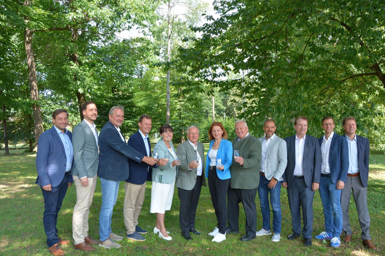 Eine Gruppe von Menschen in formeller Kleidung steht in einem Park, hält Gläser in den Händen und lächelt für ein Foto.
