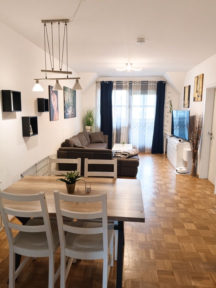 Ein modernes Wohnzimmer mit Holzboden, einer Couch, einem Esstisch und einem Fernseher. Der Raum hat weiße Wände und eine Deckenleuchte.