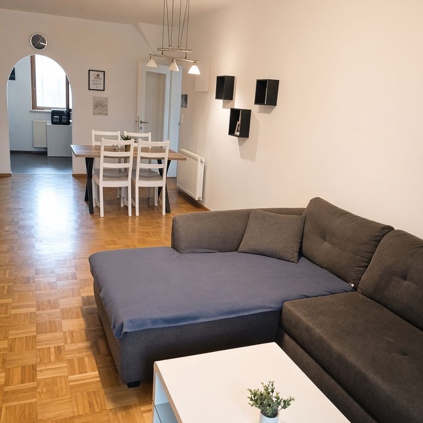 Ein modernes Wohnzimmer mit einer Couch, einem Couchtisch und einem Essbereich mit Tisch und Stühlen. Der Raum hat Holzböden und weiße Wände.