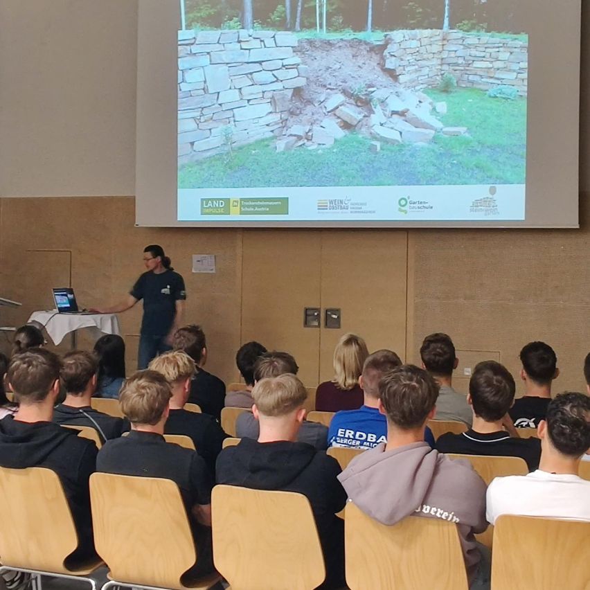 Ein Klassenzimmer mit einer Projektionsleinwand, die eine Landschaft zeigt. Eine Frau spricht zu Schülern in Stühlen.
