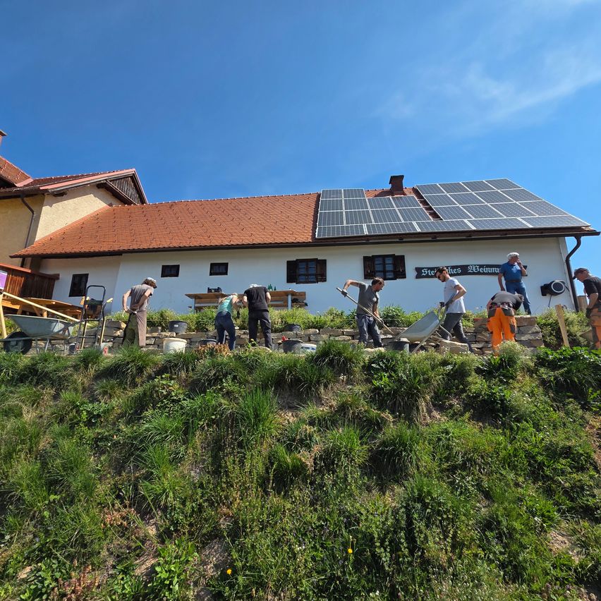 Mehrere Menschen arbeiten an einem Hang in der Nähe eines Hauses mit Solarmodulen auf dem Dach. Sie benutzen Werkzeuge wie Schaufel und Schubkarre.