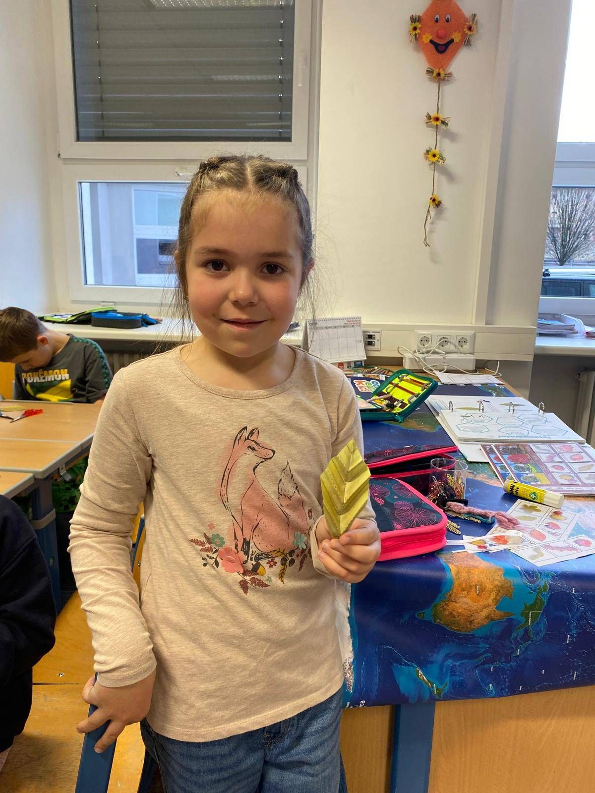 Ein junges Mädchen steht in einem Klassenzimmer und hält ein goldenes Blatt in der Hand. Hinter ihr ist ein Schreibtisch mit verschiedenen Schulutensilien übersät. Ein weiterer Schüler sitzt in der Nähe, und ein Fenster mit Jalousien ist im Hintergrund sichtbar.