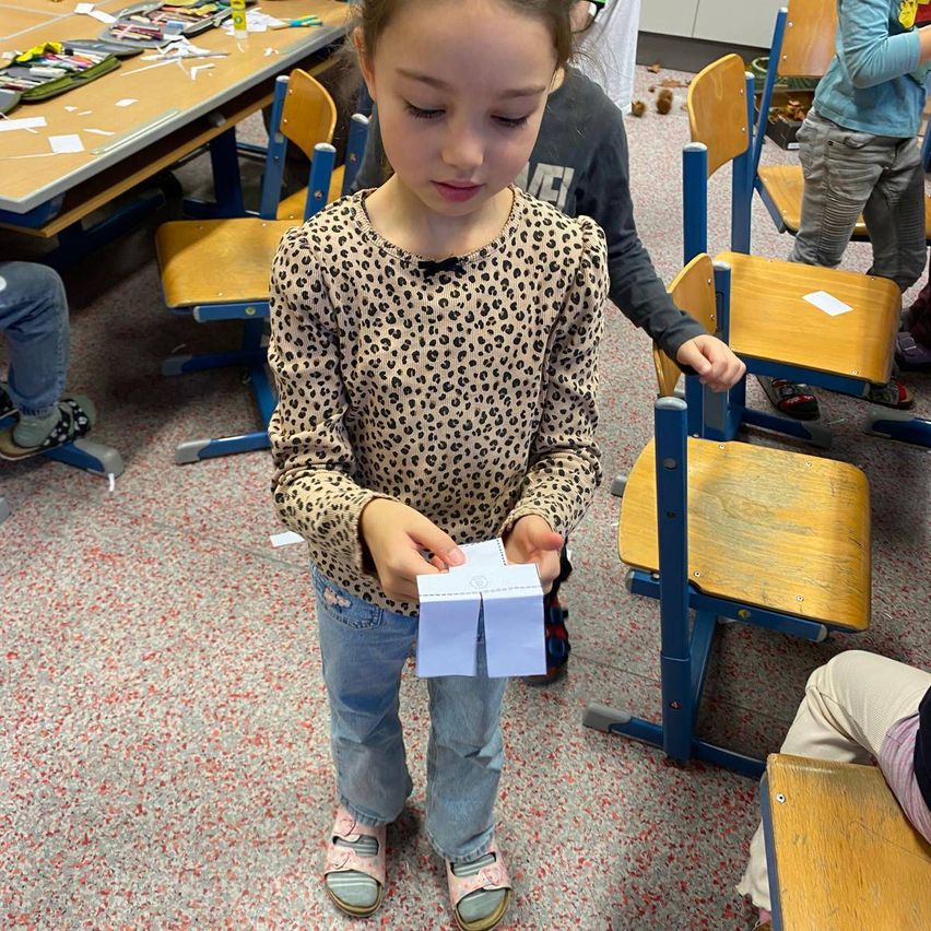 Ein kleines Mädchen mit einem Leopardenmusterhemd steht in einem Klassenzimmer und hält ein Stück Papier in der Hand, umgeben von anderen Schülern.