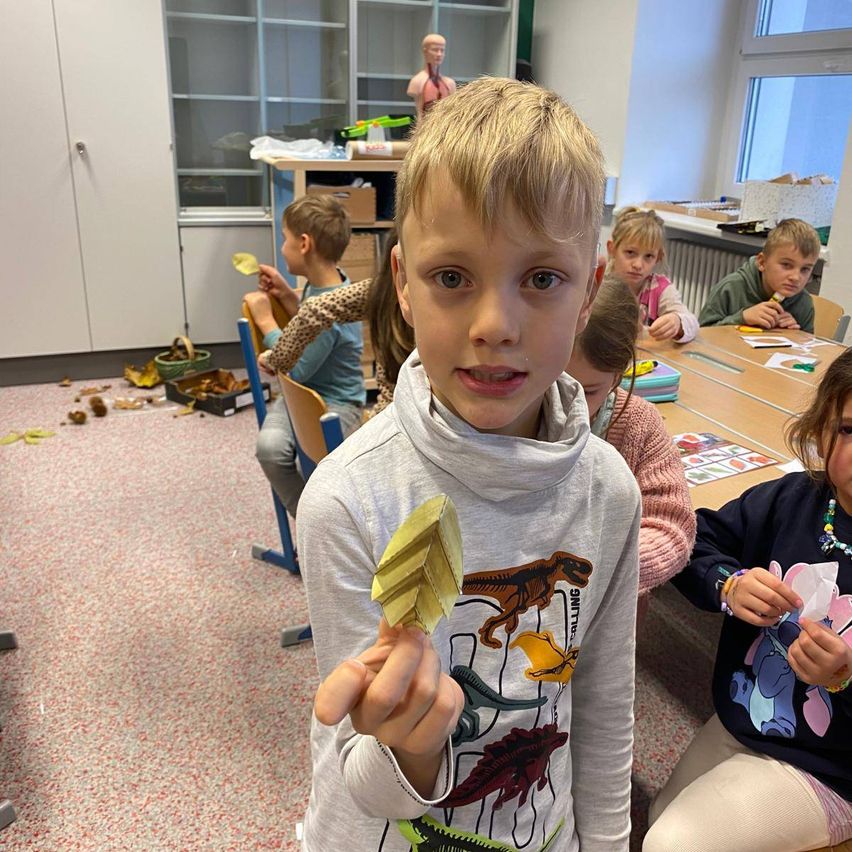 Eine Gruppe von Kindern sitzt an einem Tisch in einem Klassenzimmer und ist mit Bastelaktivitäten beschäftigt. Ein junger Junge hält eine gelbe Papierkreation, und mehrere andere arbeiten an verschiedenen Projekten.