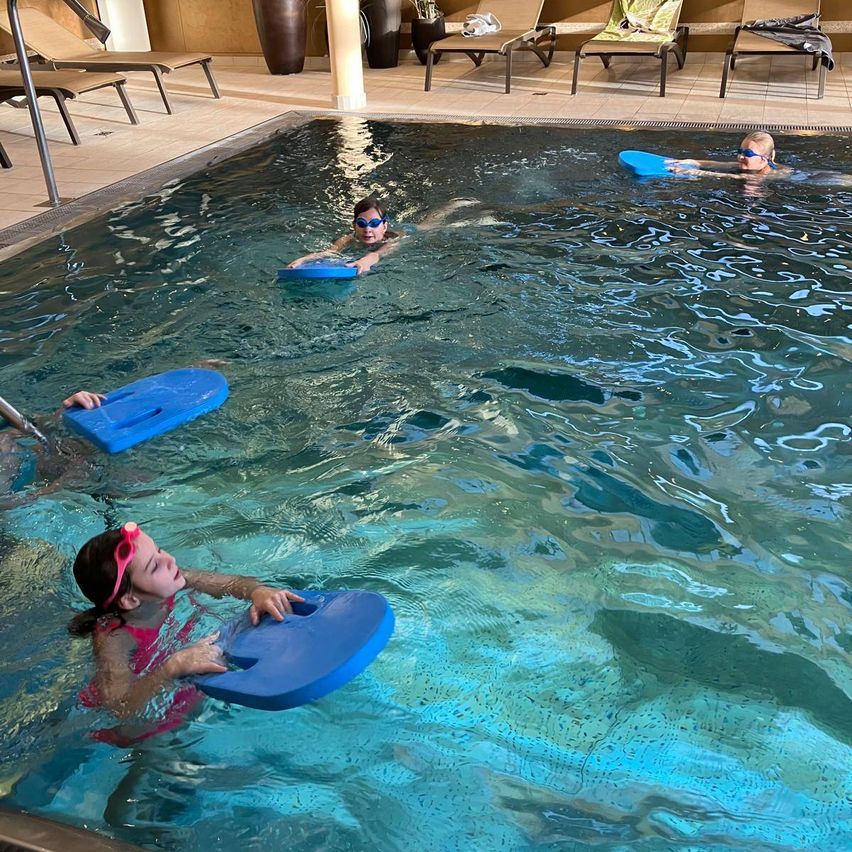 Drei Kinder in Schwimmbrillen schwimmen in einem Pool, jedes hält einen blauen Schwimmer. Der Pool ist von Liegestühlen und Topfpflanzen umgeben.