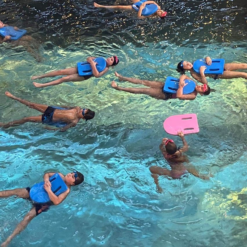 Eine Gruppe von Kindern im Schwimmbecken mit blauen Schwimmwesten und Schwimmbrillen. Ein Kind hält ein rosa Schwimmbrett. Das Wasser ist klar.