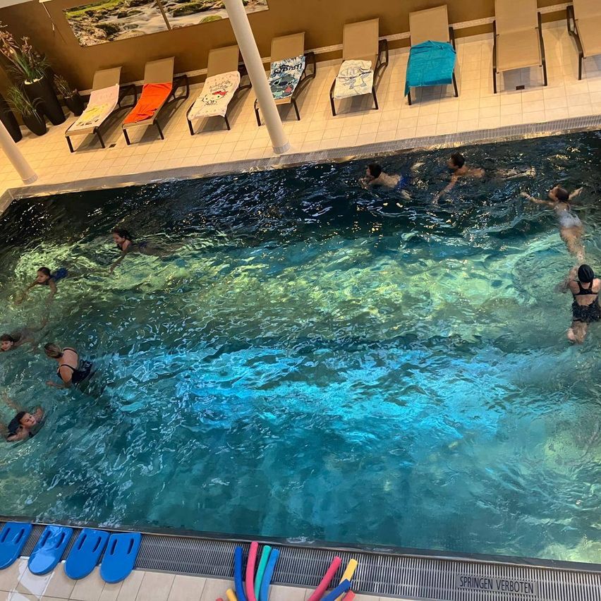 Mehrere Menschen schwimmen in einem großen Innenpool. Der Pool ist von Liegestühlen und Topfpflanzen umgeben. Der Pool hat klares blaues Wasser und wird von Deckenleuchten beleuchtet.