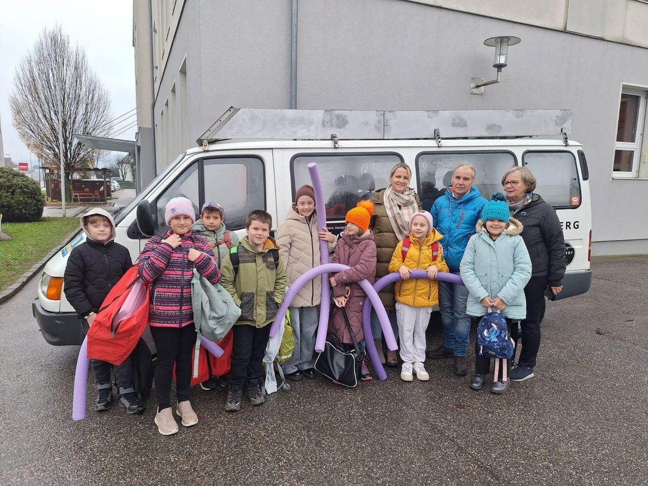 Eine Gruppe von Kindern und Erwachsenen steht lächelnd vor einem weißen Van. Sie tragen alle Winterkleidung. Die Kinder halten lila Luftballons.