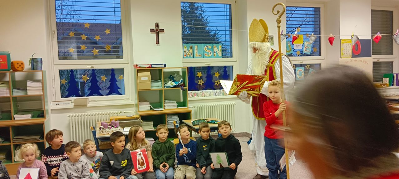 Der Heilige Nikolaus liest ein Buch für eine Gruppe von Kindern in einem Klassenzimmer vor. Sie sitzen auf dem Boden.