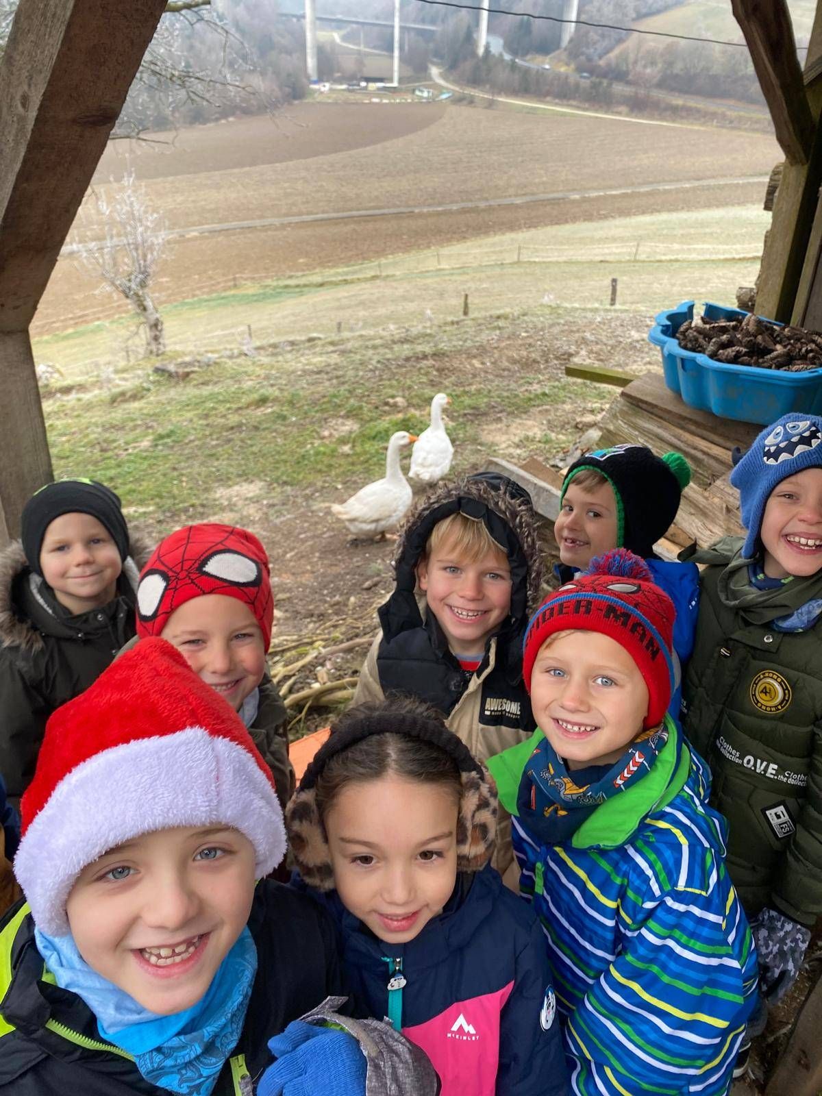 Eine Gruppe von Kindern, alle in Winterjacken und Mützen, lächeln für ein Foto. Sie sind draußen in einem ländlichen Gebiet mit einem Feld und einer Gans im Hintergrund.