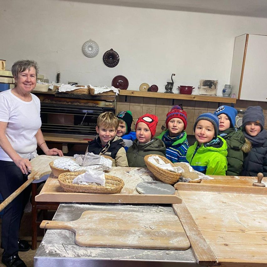 Eine Frau in einem weißen T-Shirt steht neben einem Tisch mit Teig, umgeben von Kindern in Winterkleidung, einige tragen thematische Mützen, in einer Küche mit Regalen und einem Ziegelofen.