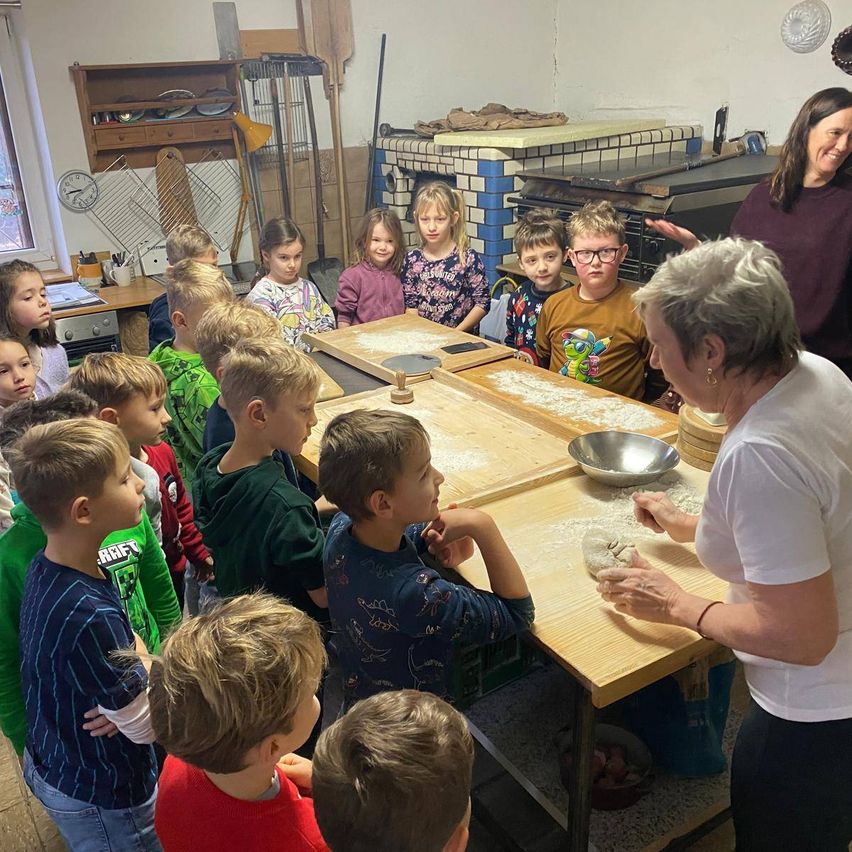 Eine Gruppe von Kindern beobachtet eine erwachsene Frau, die auf einem Tisch in einem Raum Teig herstellt.