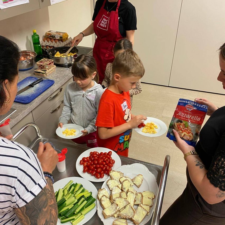 Eine Gruppe von Kindern ist in einer Küche und bereitet Essen zu. Einer hält einen Teller mit Eiern, ein anderer ein Messer. Verschiedene Zutaten sind auf Tellern, einschließlich Tomaten und Gurken. Eine Frau hält eine Packung Mozzarella.