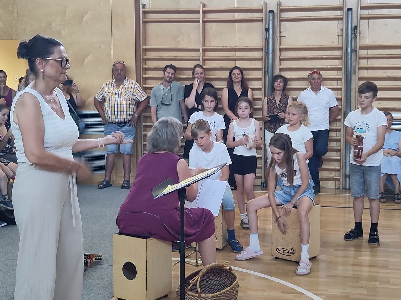 Eine Gruppe von Menschen sitzt auf Hockern und hört einer älteren Frau zu, die mit einem Notenständer steht. Dahinter stehen weitere Menschen und beobachten.