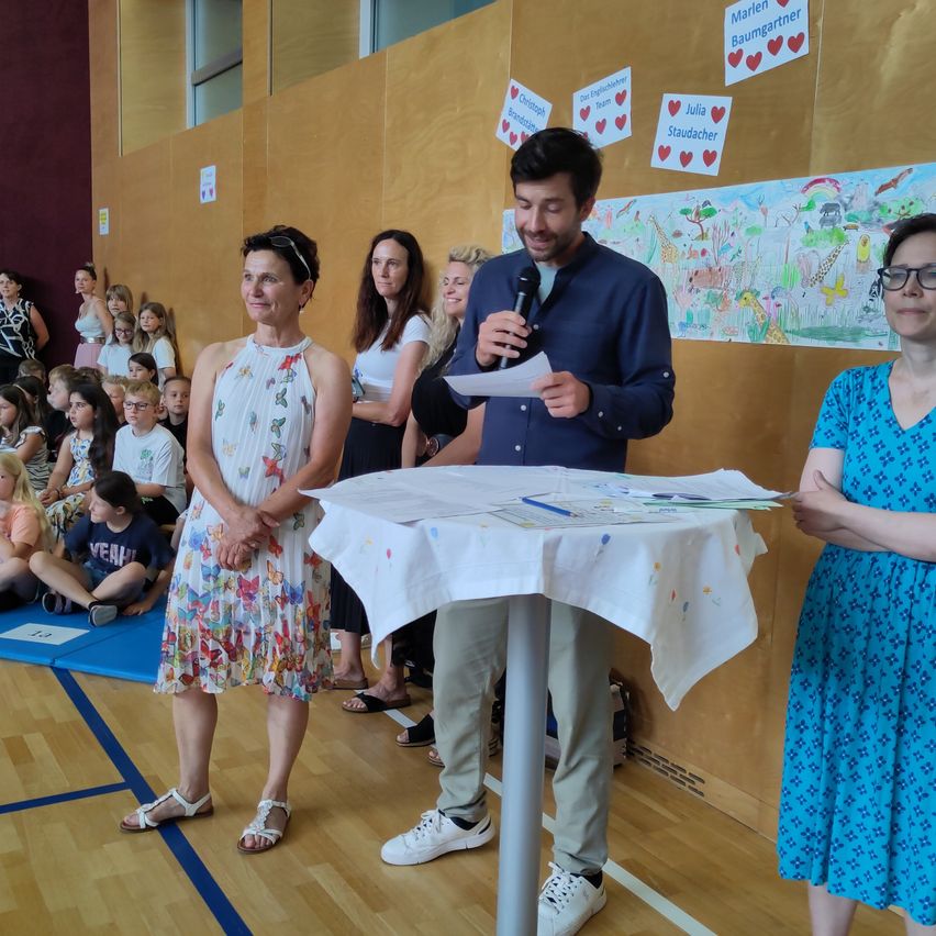 Ein Mann spricht in ein Mikrofon an einem Tisch vor einer Wand mit Kinderzeichnungen und Namen. Zwei Frauen stehen neben ihm, mit Kindern dahinter.