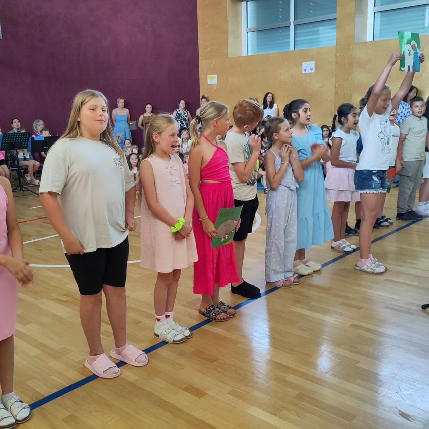 Kinder stehen in einer Reihe auf einem Basketballplatz, einige halten Bücher, tragen Freizeitkleidung und Hausschuhe. Eine Person hält ein Buch über ihren Kopf. Ein Publikum beobachtet von den Seitenlinien.
