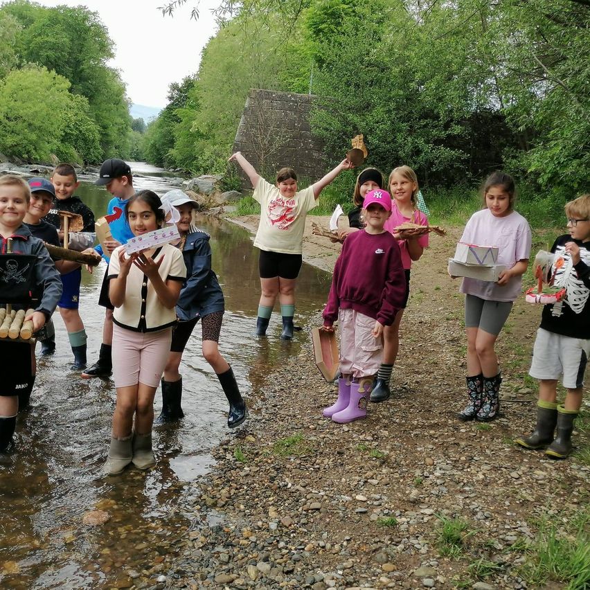 Kinder stehen in einem Fluss, halten Objekte in der Hand und tragen Stiefel. Bäume und eine Steinbrücke im Hintergrund.