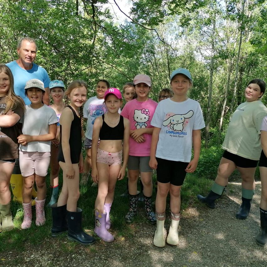 Eine Gruppe von Kindern und Erwachsenen steht in einem Wald. Die Kinder tragen Stiefel und Mützen, während die Erwachsenen T-Shirts tragen. Bäume und Pflanzen sind im Hintergrund.