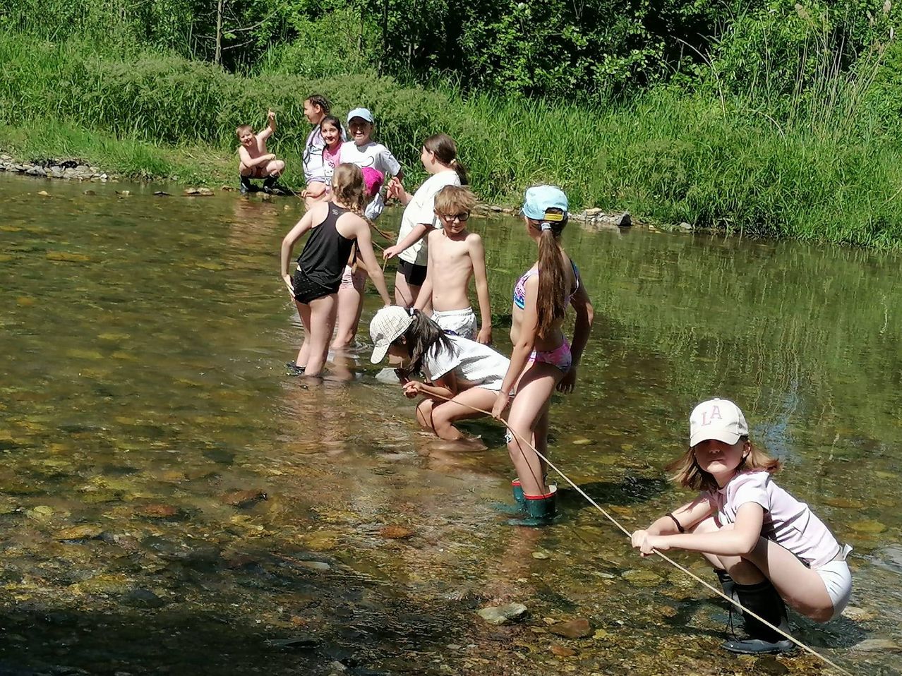 Eine Gruppe von Kindern watet in einem Fluss, trägt Mützen und Stiefel. Sie halten einen Stock, um Fische zu fangen.