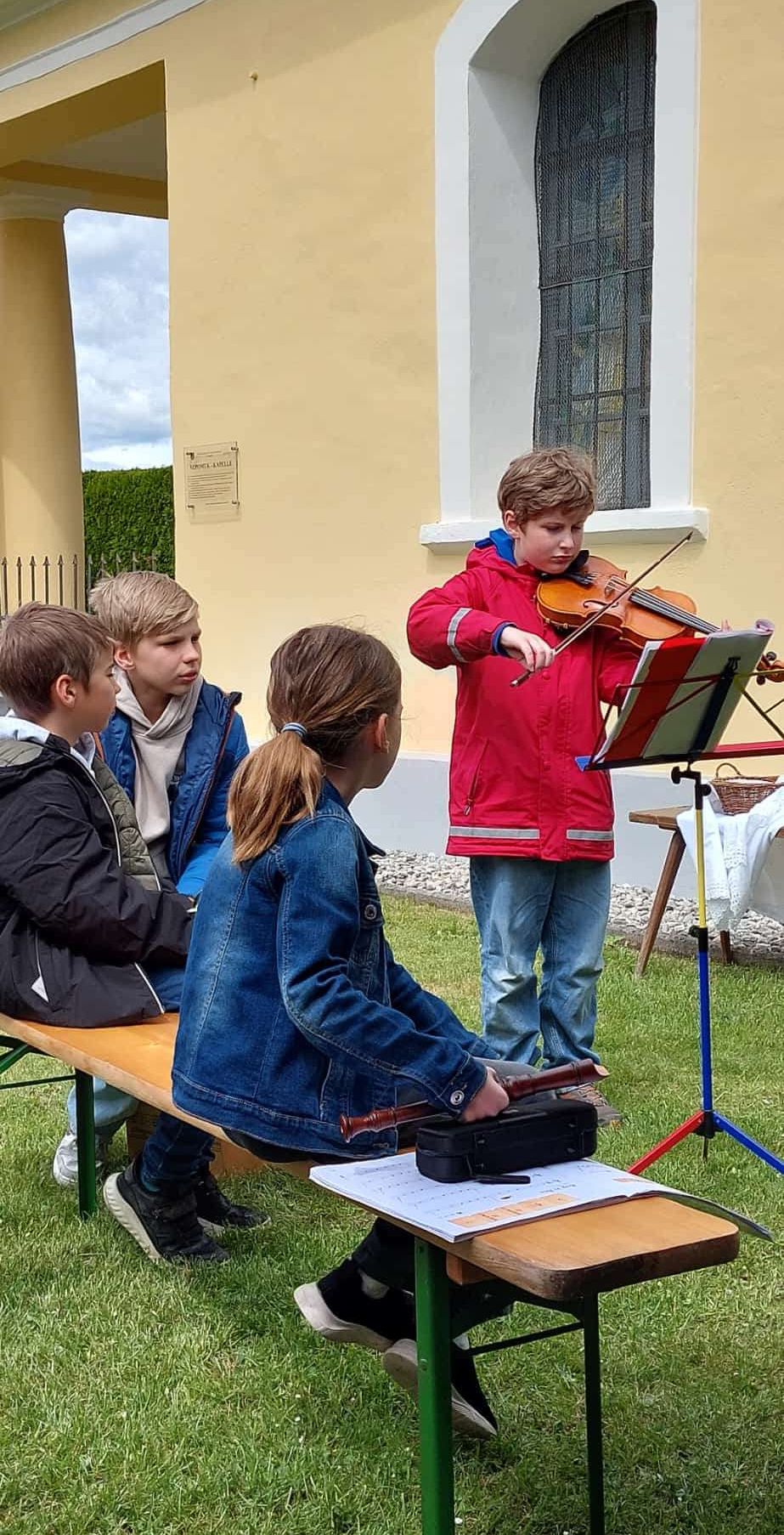 Ein Junge spielt Geige mit Notenständer vor einem Gebäude, während andere zuschauen.