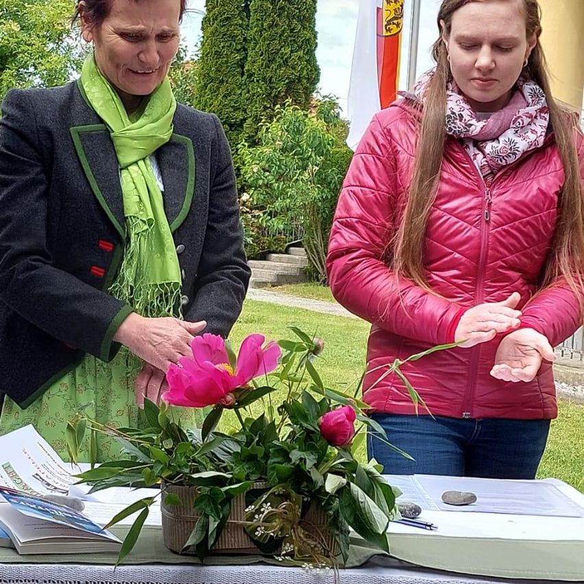 Zwei Frauen stehen neben einem Tisch mit einer Blumenarrangement, eine mit einem grünen Schal, die andere in einer roten Jacke. Sie schauen beide auf die Blumen hinunter.