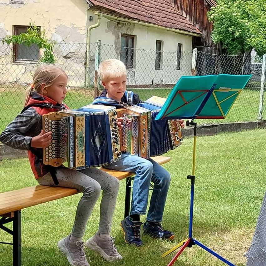 Zwei Kinder sitzen auf einer Bank in einem Rasengarten und spielen Akkordeons. Hinter ihnen steht ein Haus mit einem roten Dach und einem Zaun.