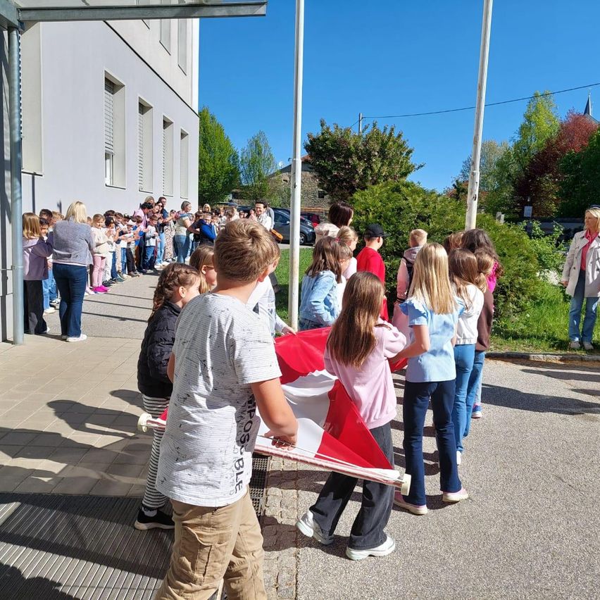 Eine Menschenmenge aus Kindern und Erwachsenen versammelt sich vor einem Gebäude und hält eine rot-weiße Fahne. Sie stehen auf einem Bürgersteig mit einem Metallgitter.