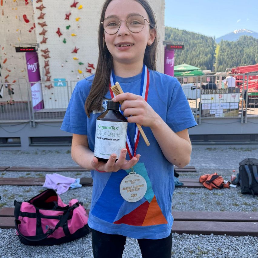 Ein Mädchen hält eine Flasche OrganoTex BioCare Woll- und Daunenwaschmittel und trägt eine Medaille und ein blaues Shirt vor einer Kletterwand.